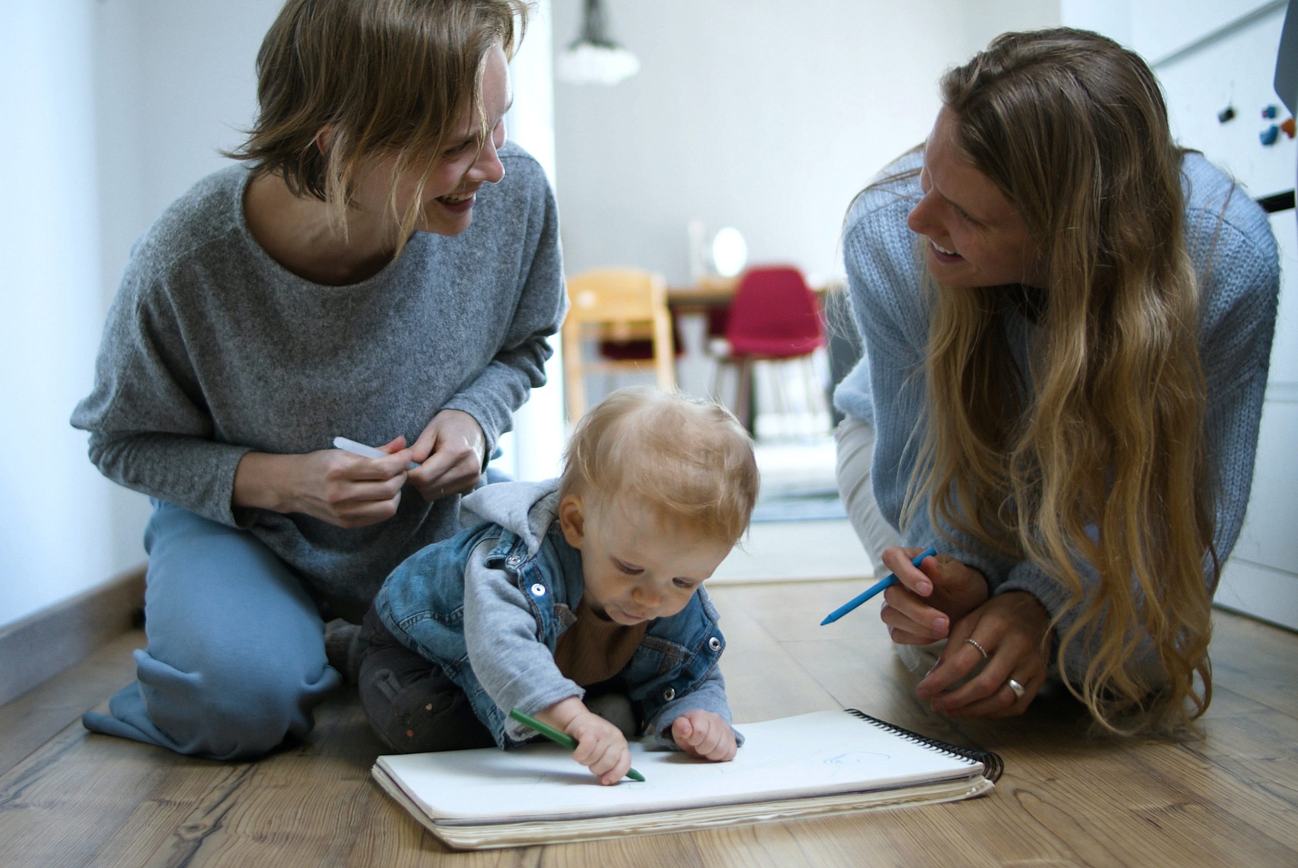 Two women kneel on the floor and smile at each other while a toddler draws on paper with a crayon